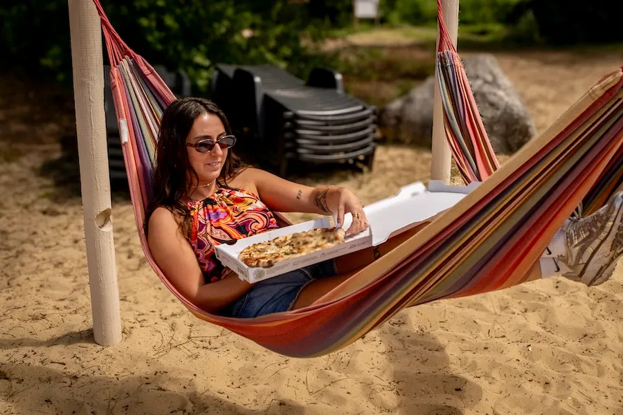  Vacancière installée dans un hamac sur la plage d'un camping, dégustant une pizza dans sa boîte