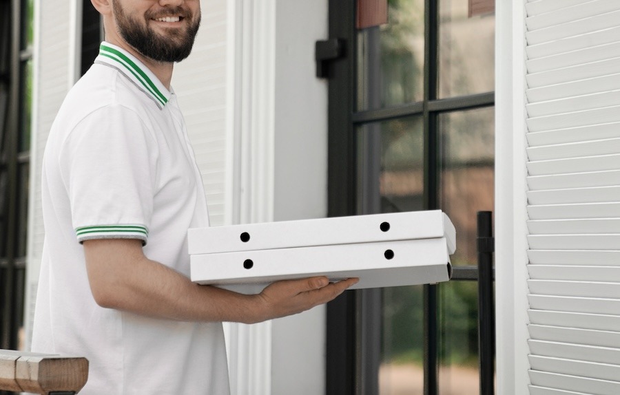 Livreur souriant remettant deux boîtes à pizza à l'entrée d'un emplacement, illustrant le service de livraison proposé aux vacanciers en camping